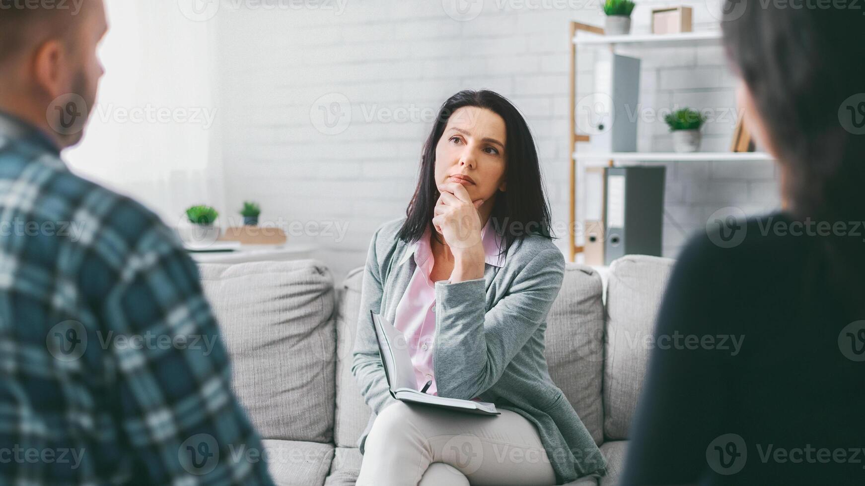 A woman sits on a couch, thoughtfully listening to two people as she holds a notepad in her lap. She appears to be a therapist, counselor, or mediator, engaged in a session with two clients. photo