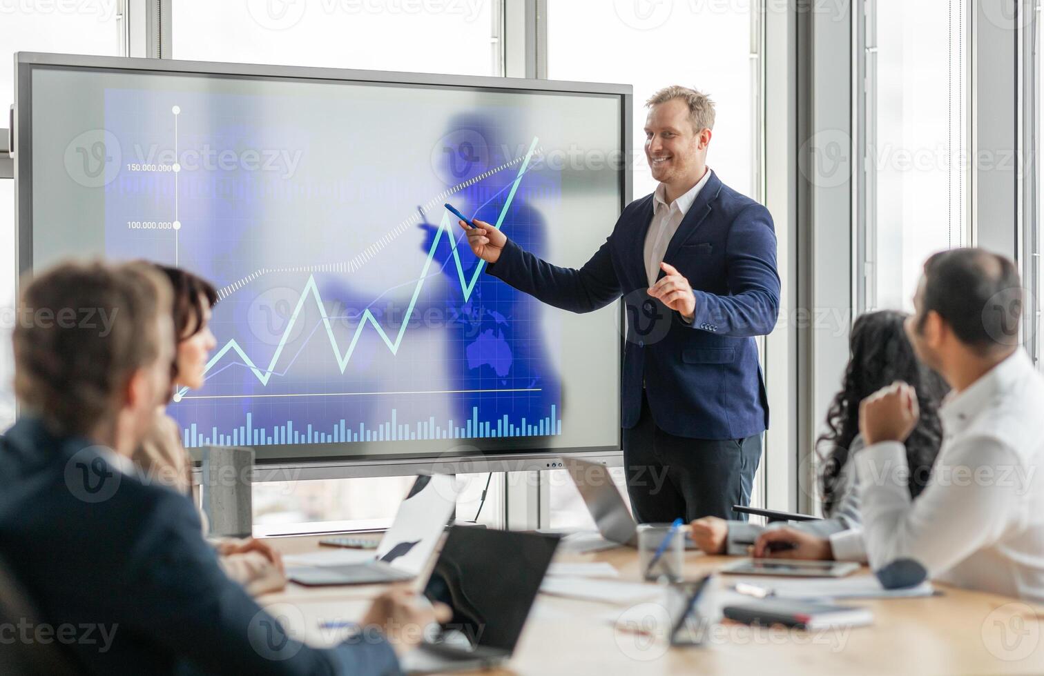 A businessman stands at the front of a meeting room, pointing at a large screen displaying a financial graph. He is presenting data to his colleagues, who are seated around a conference table. photo