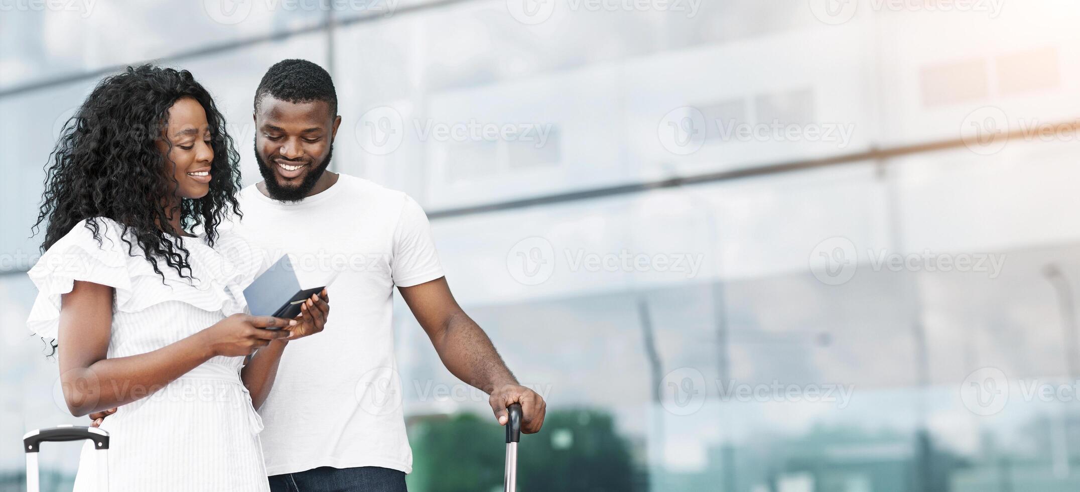 African American couple stands in front of a large glass window at an airport terminal, checking their boarding pass. They appear to be ready to travel and are smiling, copy space photo