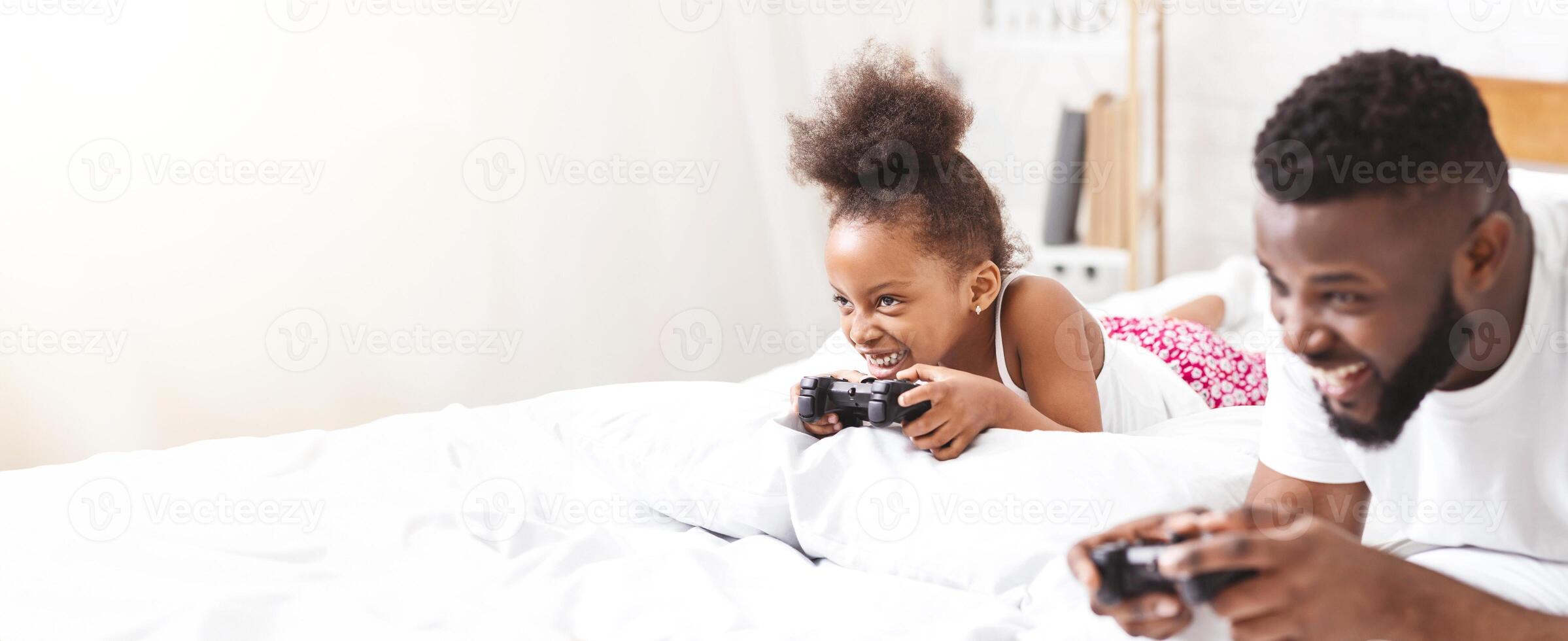 African American father and his daughter lie in bed together, both engrossed in playing games with handheld controllers. They appear to be having a lot of fun together photo