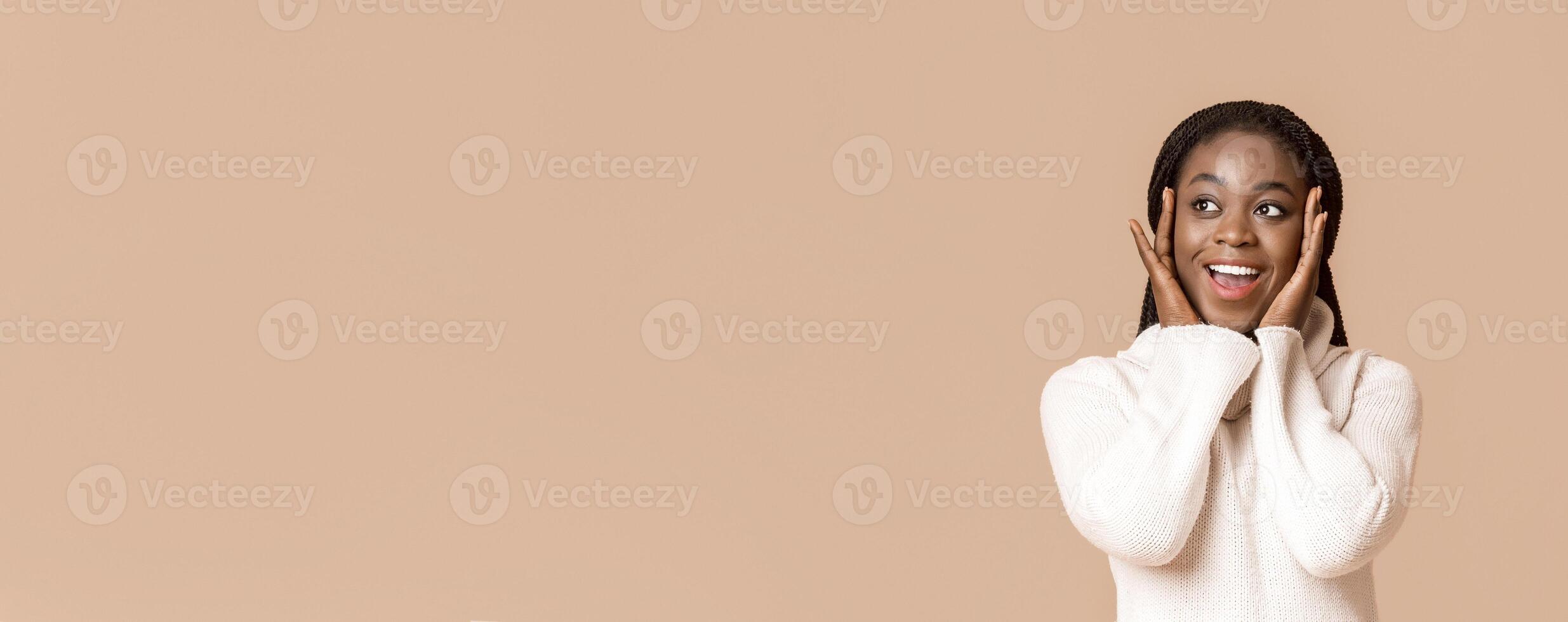 Amazed african american woman with open mouth touching her cheeks with hands over beige background, looking at empty space, panorama photo