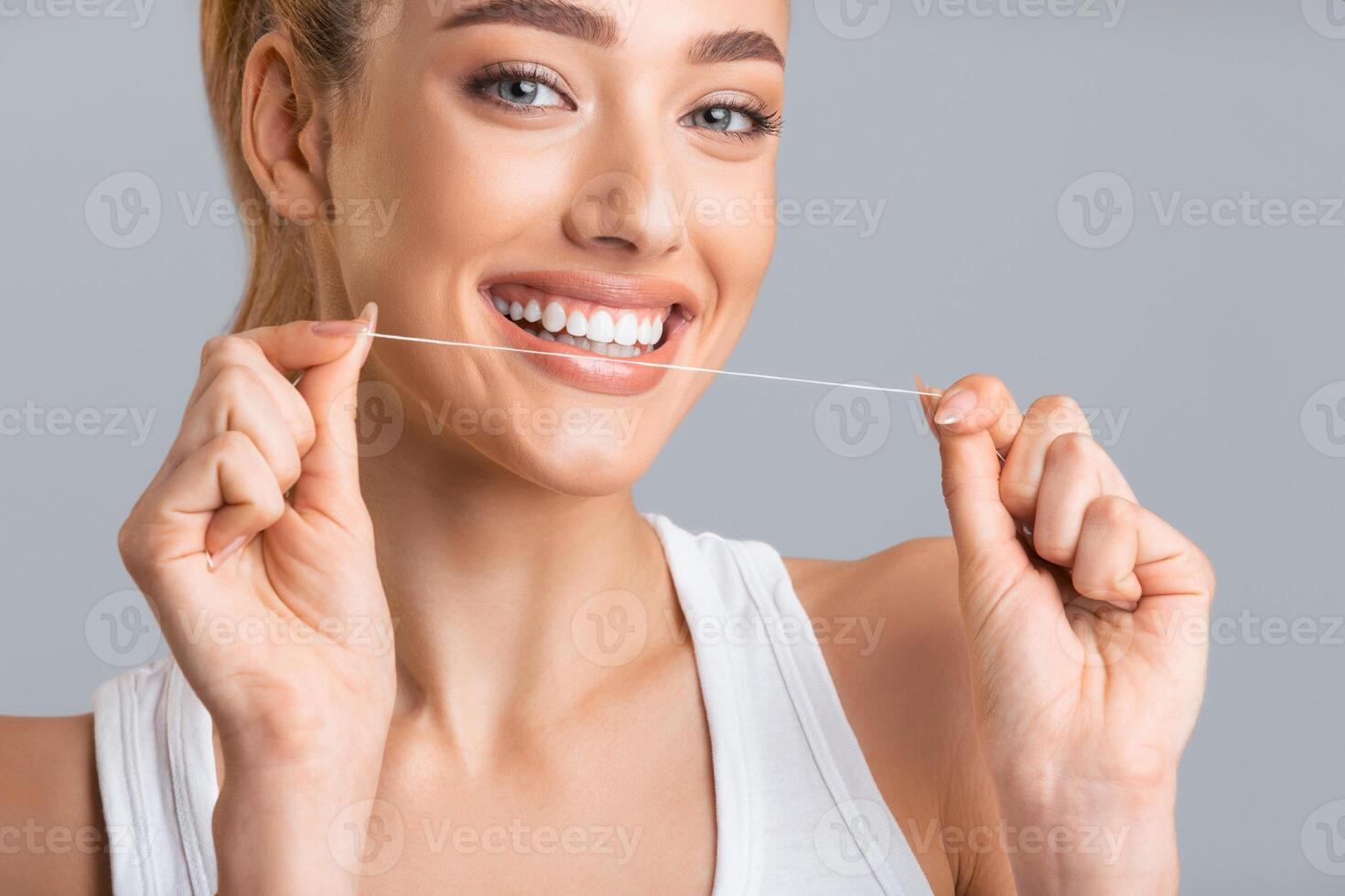 A young woman with long blonde hair is flossing her teeth with a white floss. She is smiling and looking at the camera. The background is a solid grey. photo