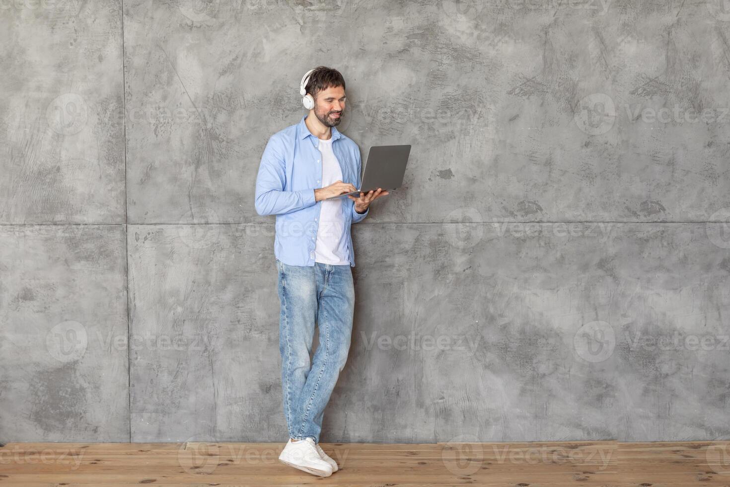 A man in a blue shirt, white t-shirt, and jeans stands in front of a concrete wall and uses a laptop computer. He is wearing headphones and smiling. He is standing on a wooden floor. photo