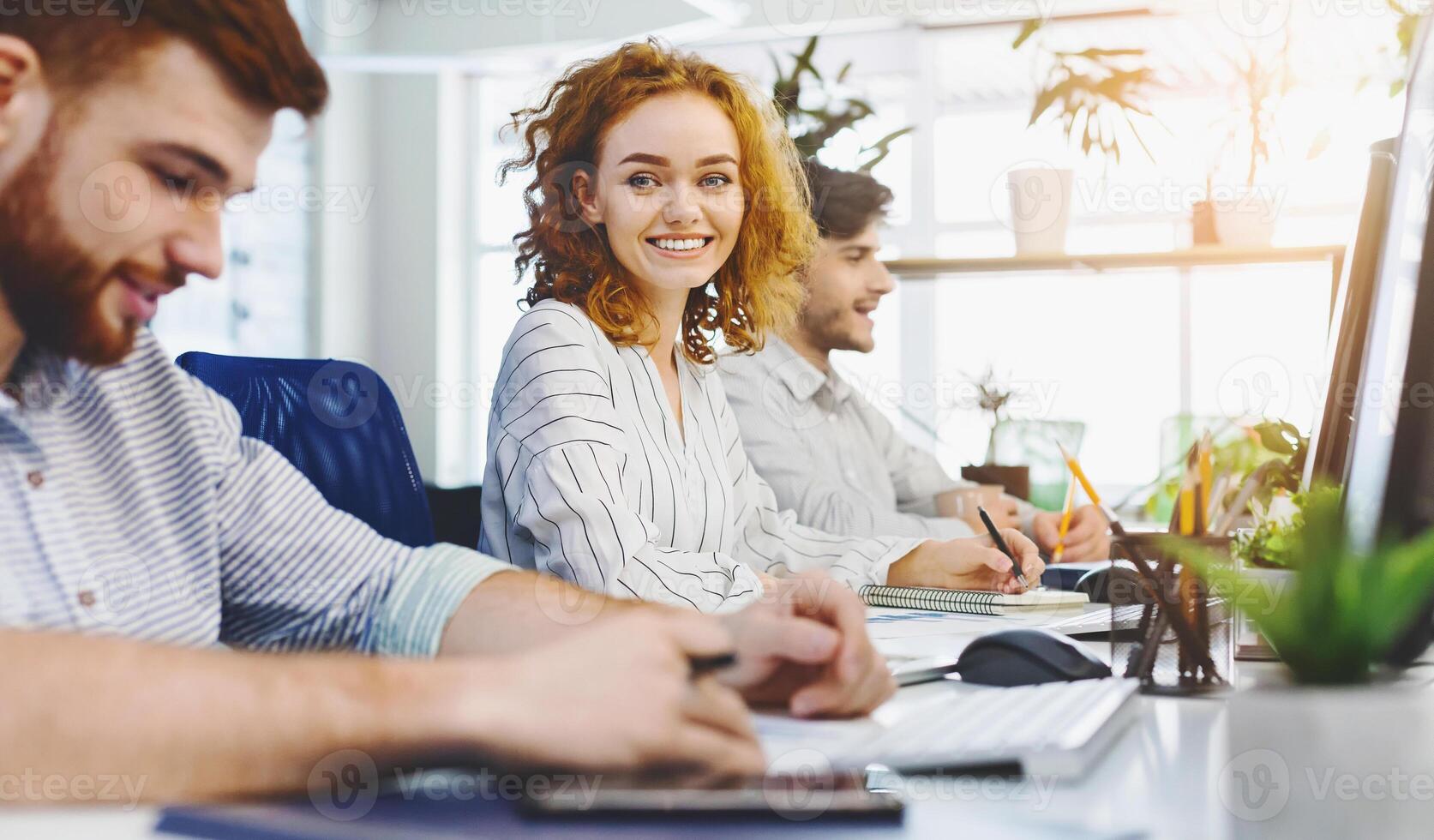 Cheerful redhead girl smiling at camera, working at coworking with other people, empty space photo