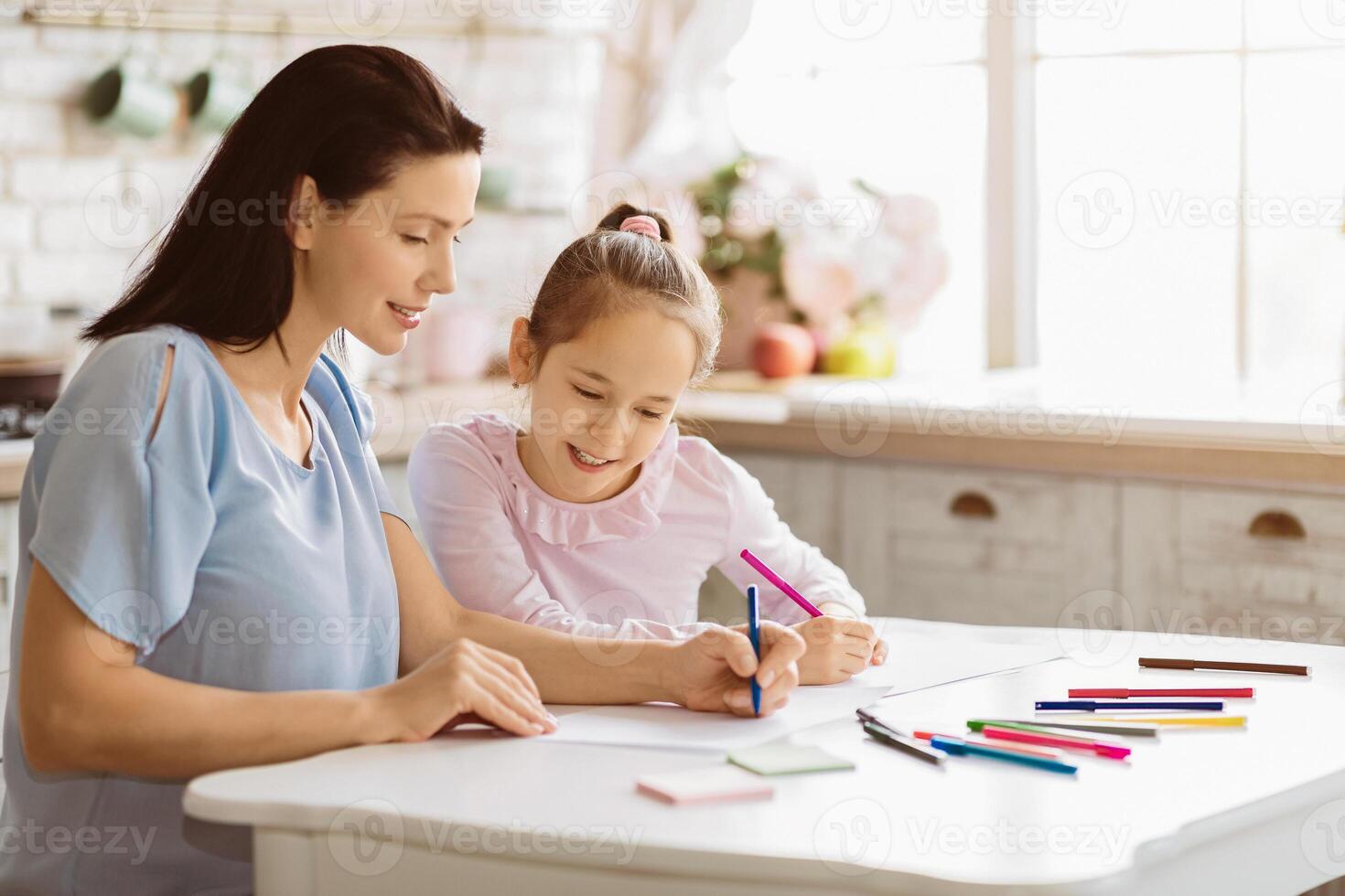 A young girl sits at a table in a kitchen with her mother, both drawing with colored pencils. The mother is watching the girl and smiling. photo