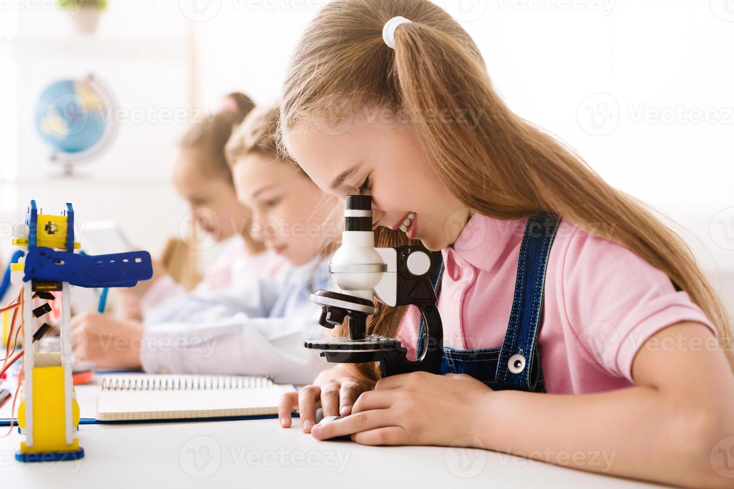 A young girl sits at a desk in a classroom, peering into a microscope, while other students sit beside her. The image highlights the importance of hands-on learning in STEM education for children. photo