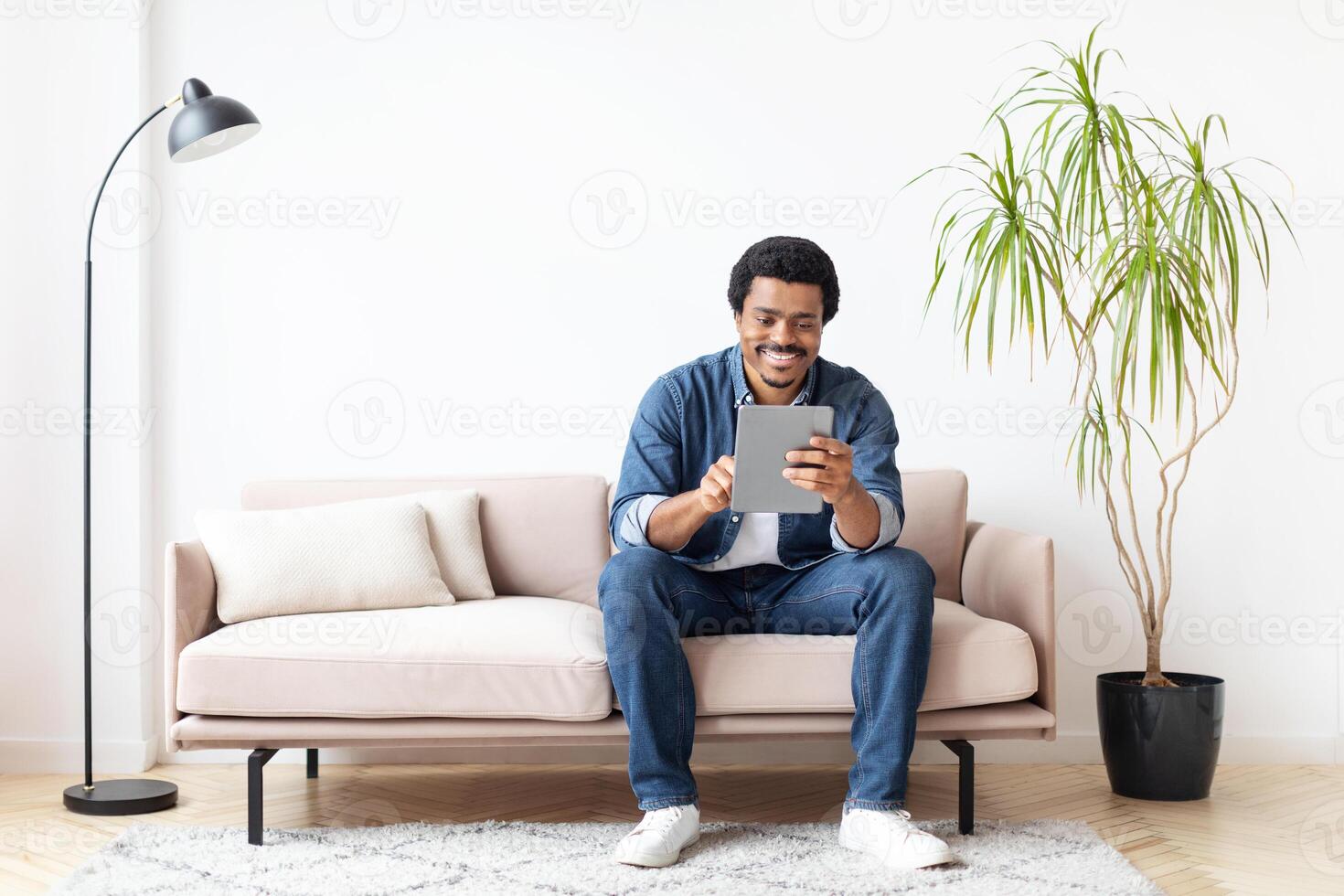 African American man is seated on a couch engaging with a tablet device in this indoor setting, showcasing modern technology and leisure in a home environment. photo