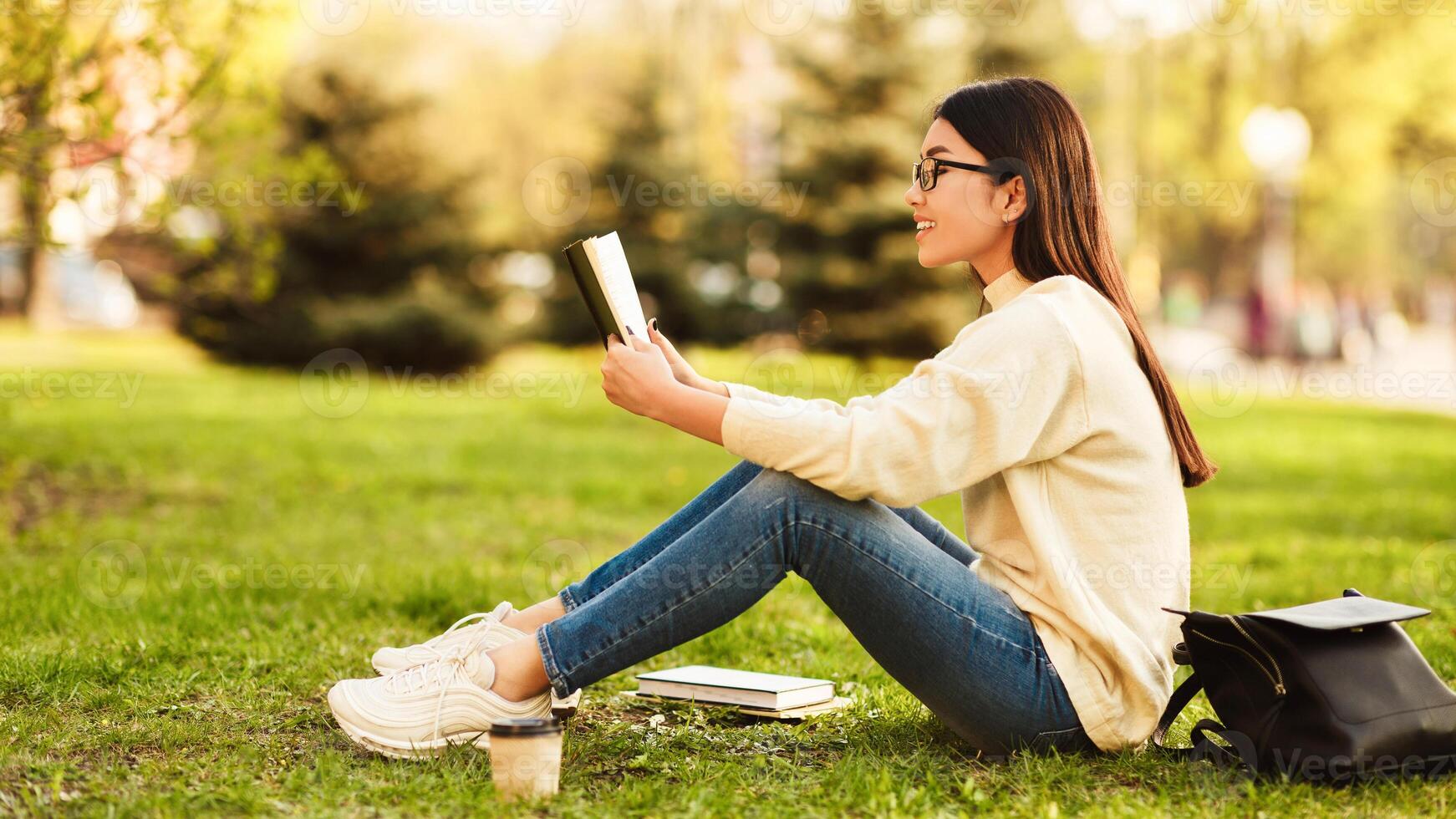 Asian woman student is seated on the green grass, engrossed in reading a book. She is alone, focused on the pages in front of her, with a serene expression on her face. photo