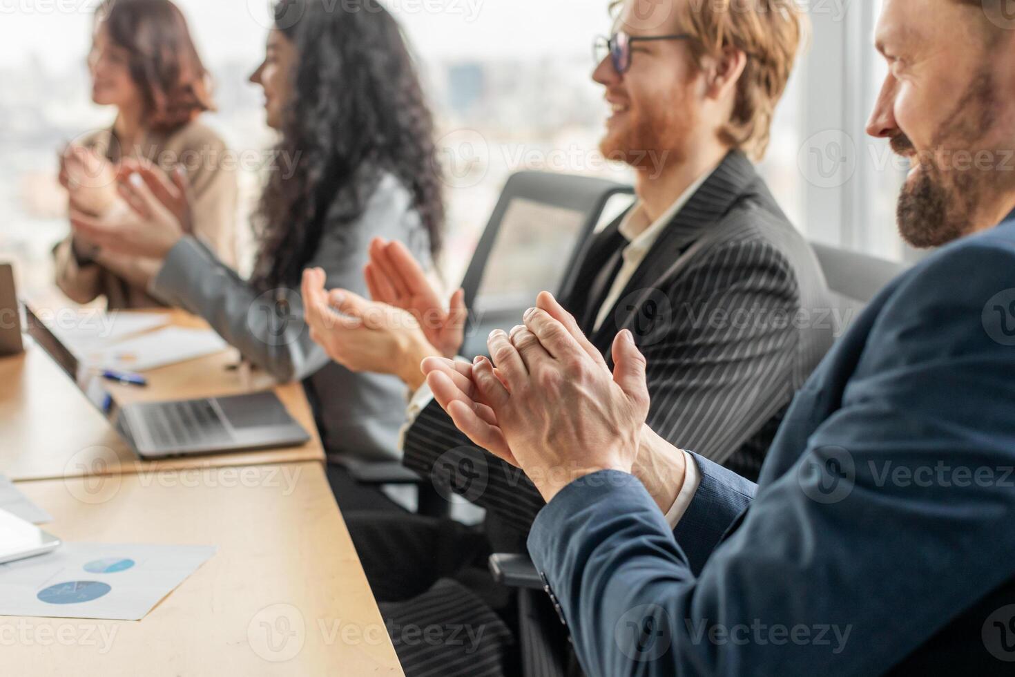 A group of business professionals are seated around a conference table, clapping their hands in appreciation. A laptop and papers are visible on the table. photo