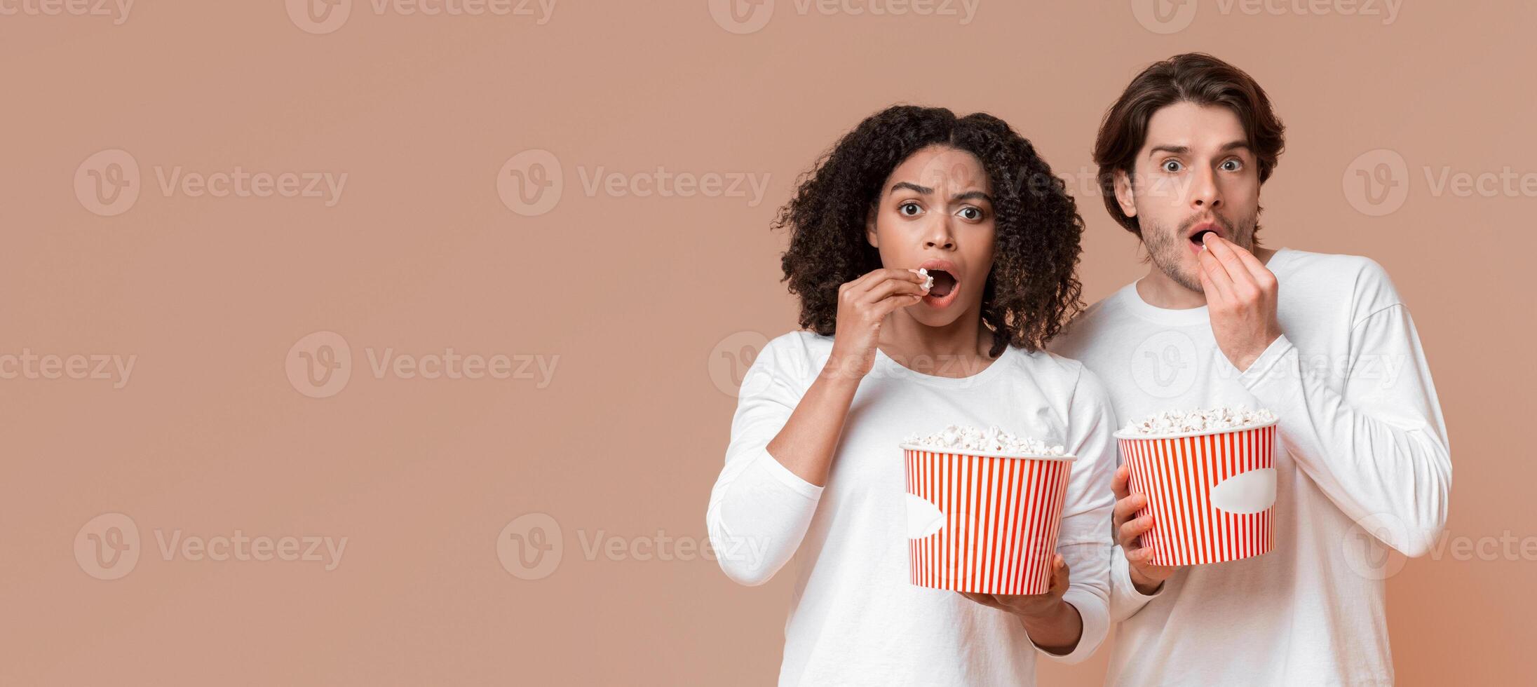 Scary movie. Shocked multiracial couple eating popcorn from buckets and looking at camera with scared face expression, yellow background photo