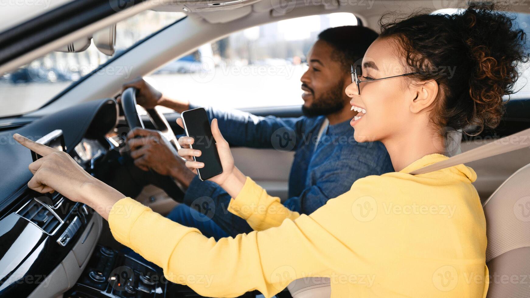 A young black woman is sitting in the passenger seat of a car. She is using her phone, likely a navigation app, as she points to the screen and smiles. The driver, a man, is looking straight ahead photo