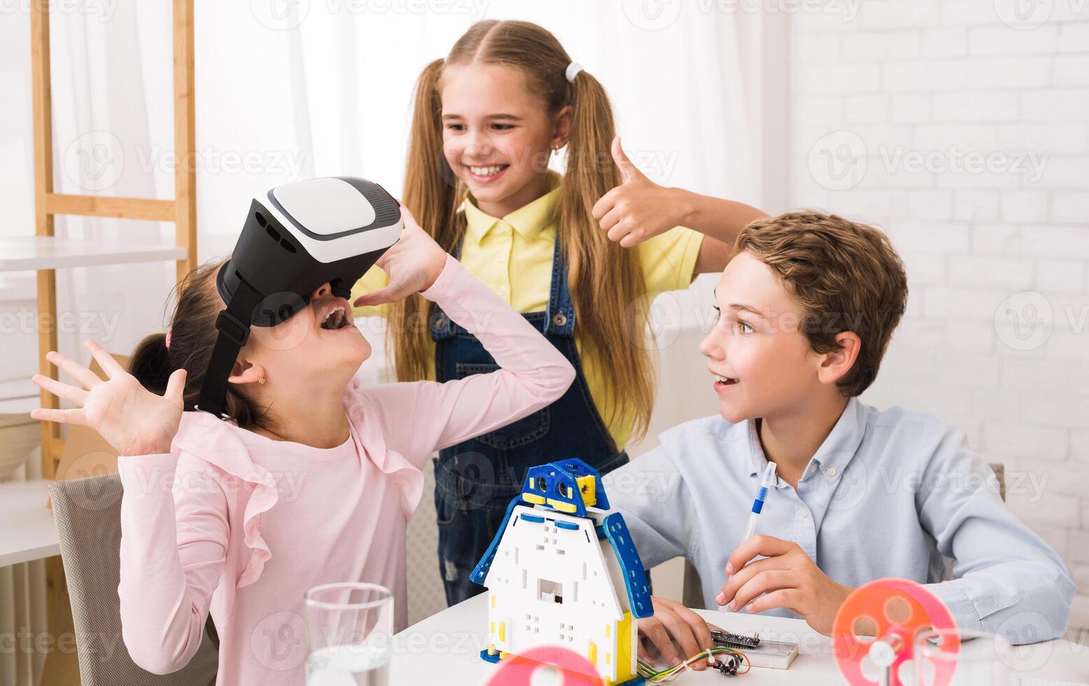Three children are working on a robotics project. One girl is wearing a virtual reality headset and has a look of excitement on her face. The other two children are watching her, photo