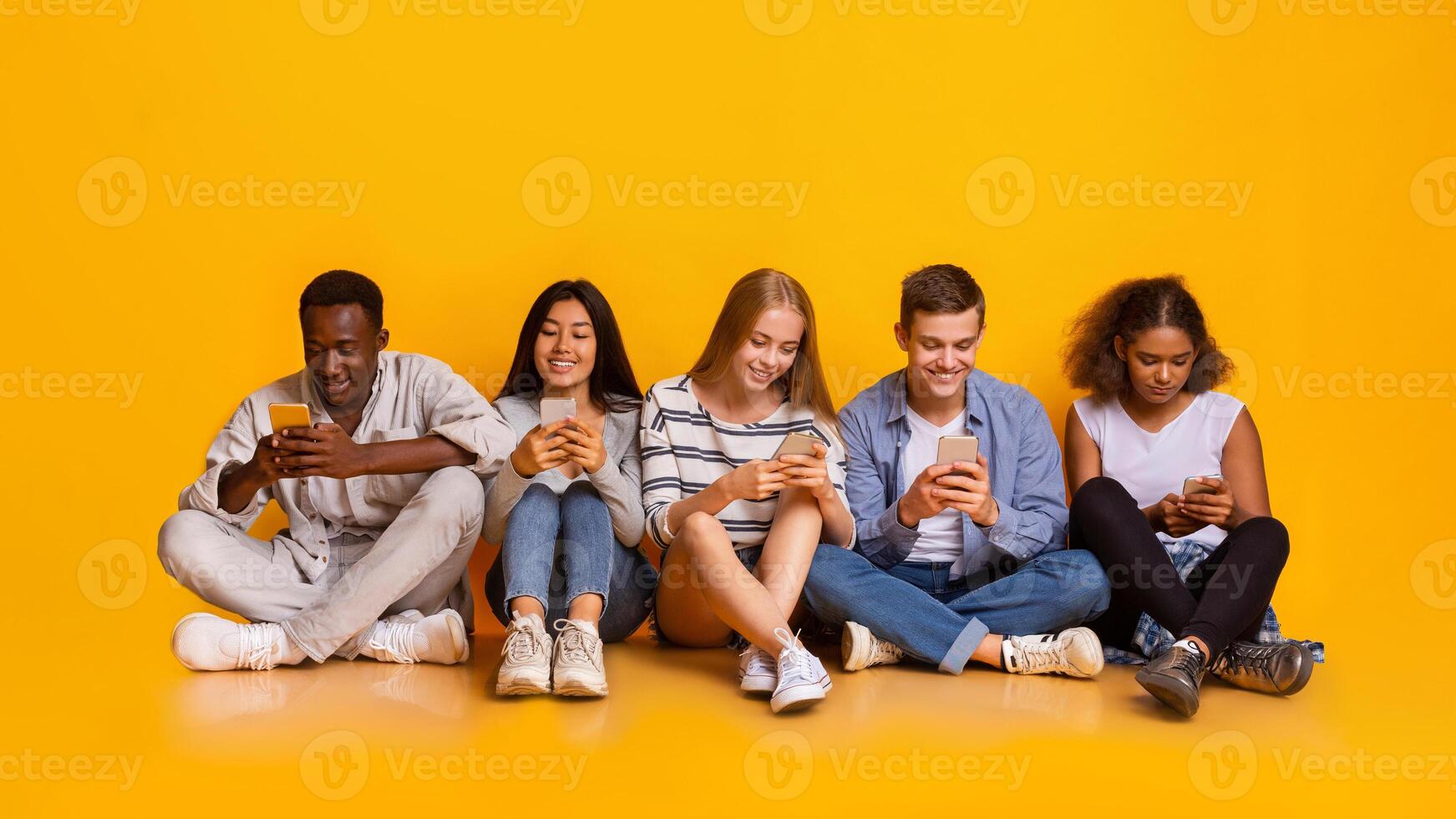 Multiracial teenagers sitting on floor and texting with their smartphones, yellow background, concept about technology and global communication photo