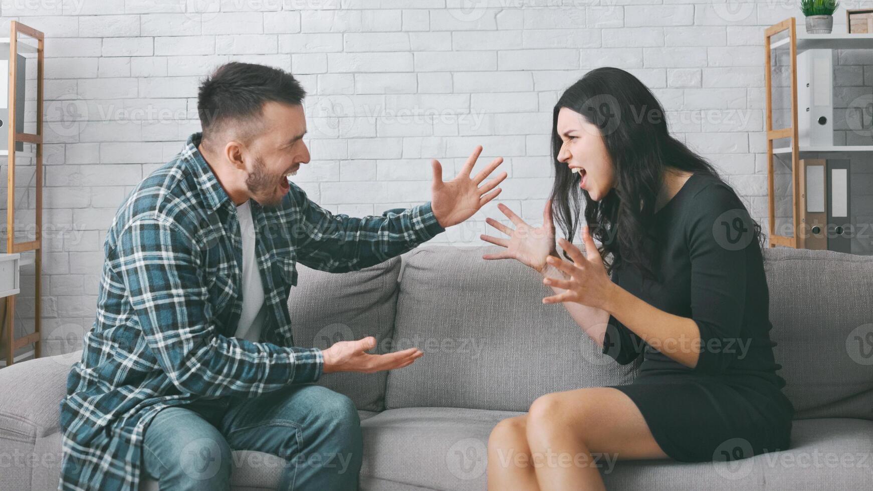 A couple is having a heated argument on a couch in their living room. The woman is sitting on the couch with her legs crossed and is gesturing wildly with her hands while yelling at the man. photo