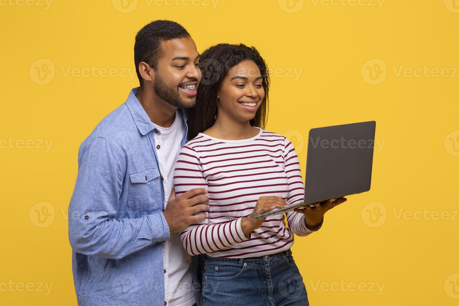 Black man and woman standing side by side, both looking intently at a laptop screen in front of them. They appear focused and engaged in whatever content is being displayed on the computer. photo