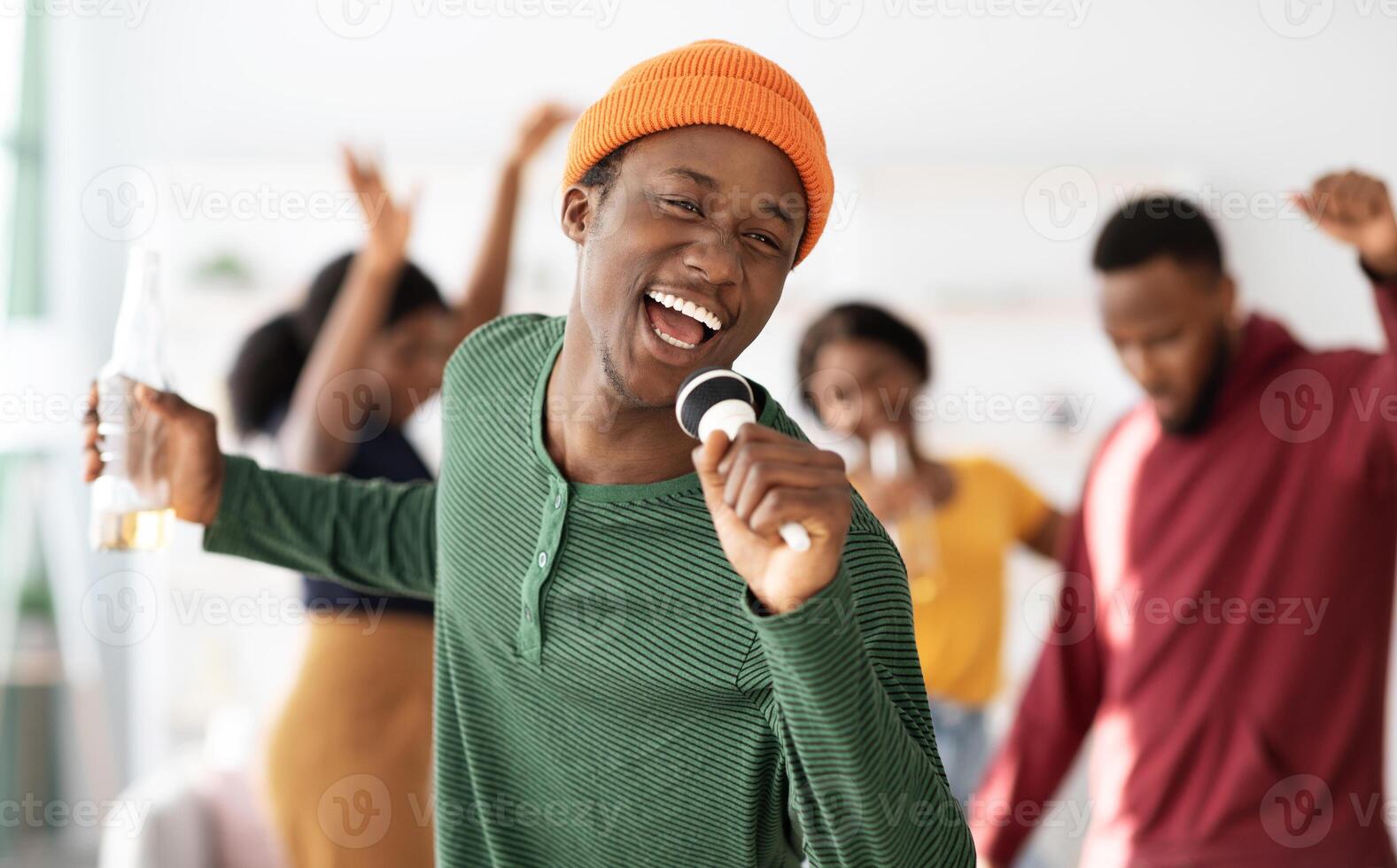 Positive african american young man with bottle of beer singing karaoke while having home party with friends, holding microphone and singing songs, selective focus, blurred background photo