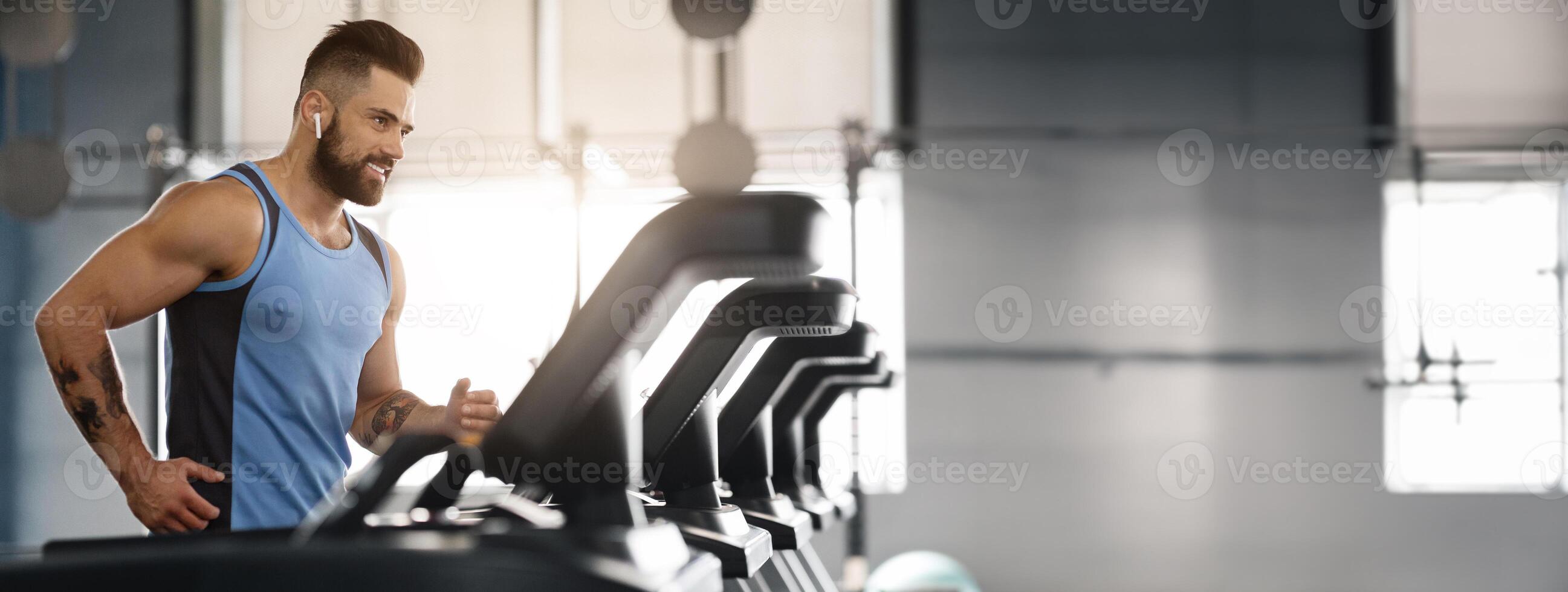 Jogging with pleasure. Young handsome man in modern wireless headphones smiling, running on treadmill at gym, free space photo