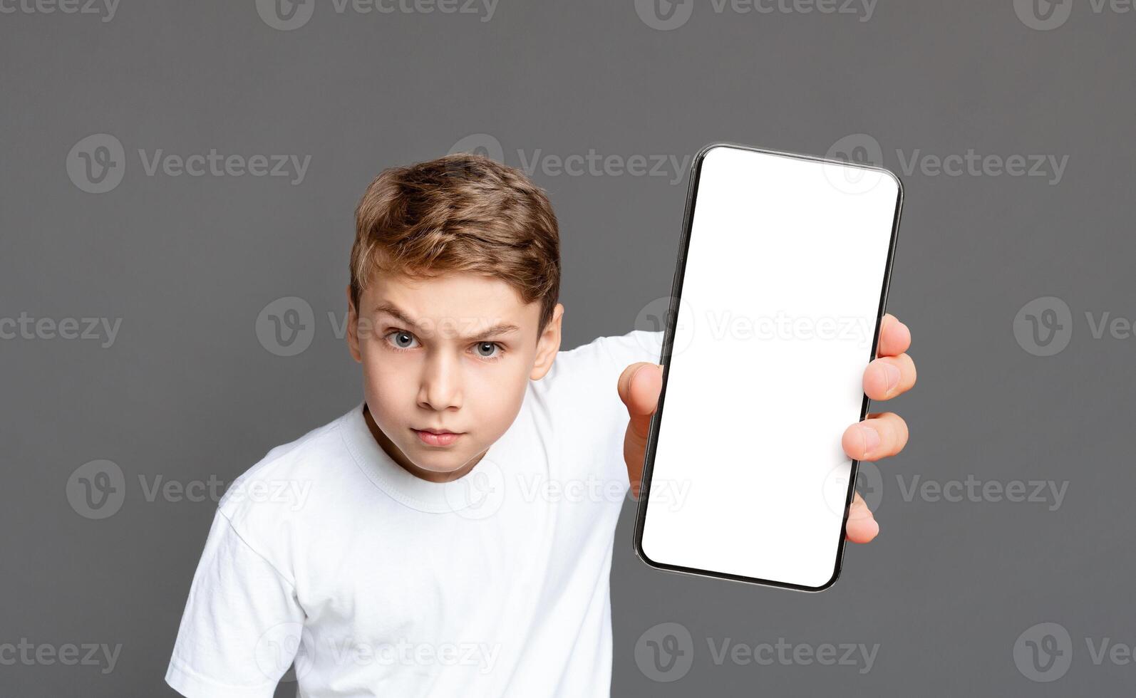 A young boy with a serious expression holds out a smartphone with a blank screen towards the camera on a gray background photo