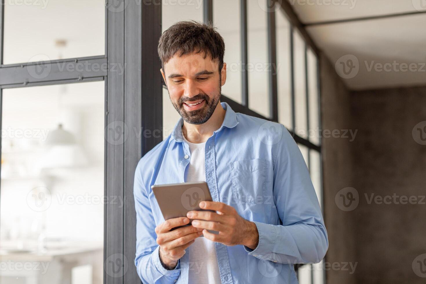 A man in a blue shirt stands in an office building, using a tablet device while looking at the screen with a smile. The man is in a casual work environment, surrounded by large windows. photo