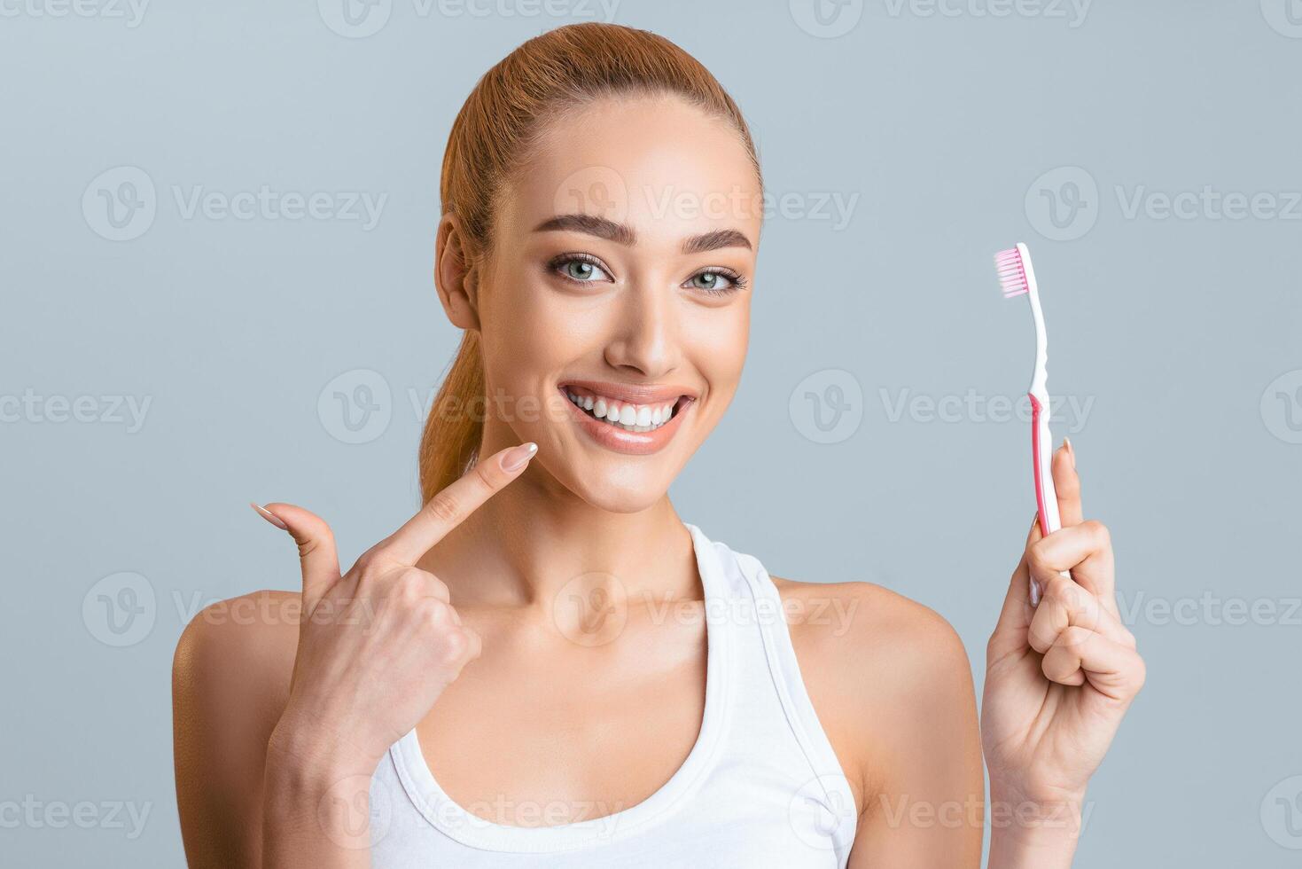 Woman in a white tank top smiling brightly as she points at her teeth with her left hand and holds a toothbrush in her right hand. She appears to be happy and confident about her teeth. photo