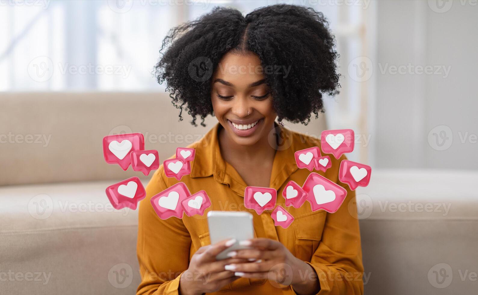 A young black woman with curly hair is sitting at home and smiling while checking her smartphone. She is surrounded by pink heart icons, indicating that she is receiving many likes photo