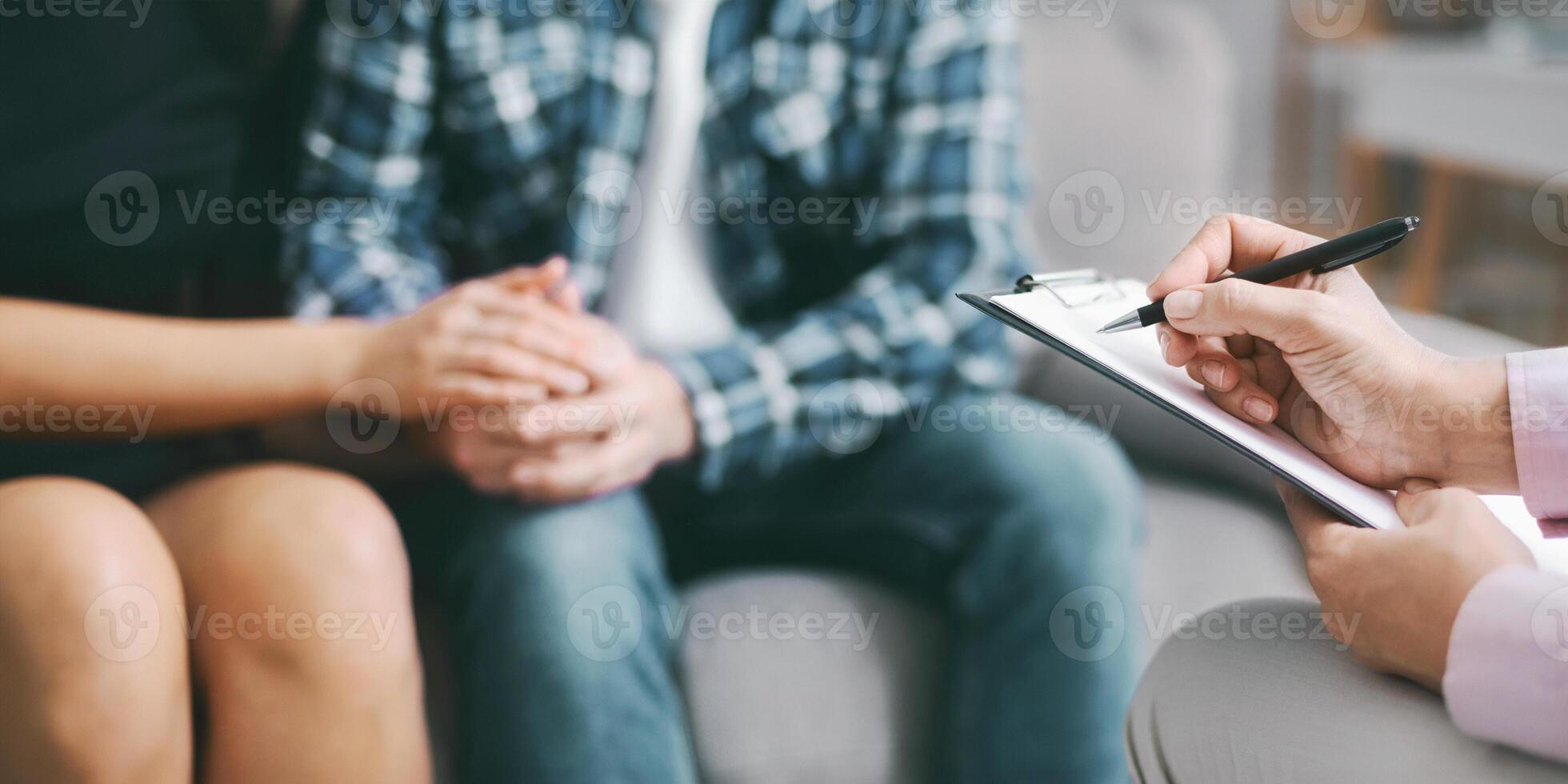 The image shows a therapist taking notes on a clipboard with a pen, while a couple sits together in a counseling session. They are holding hands, suggesting a close connection and shared experience photo