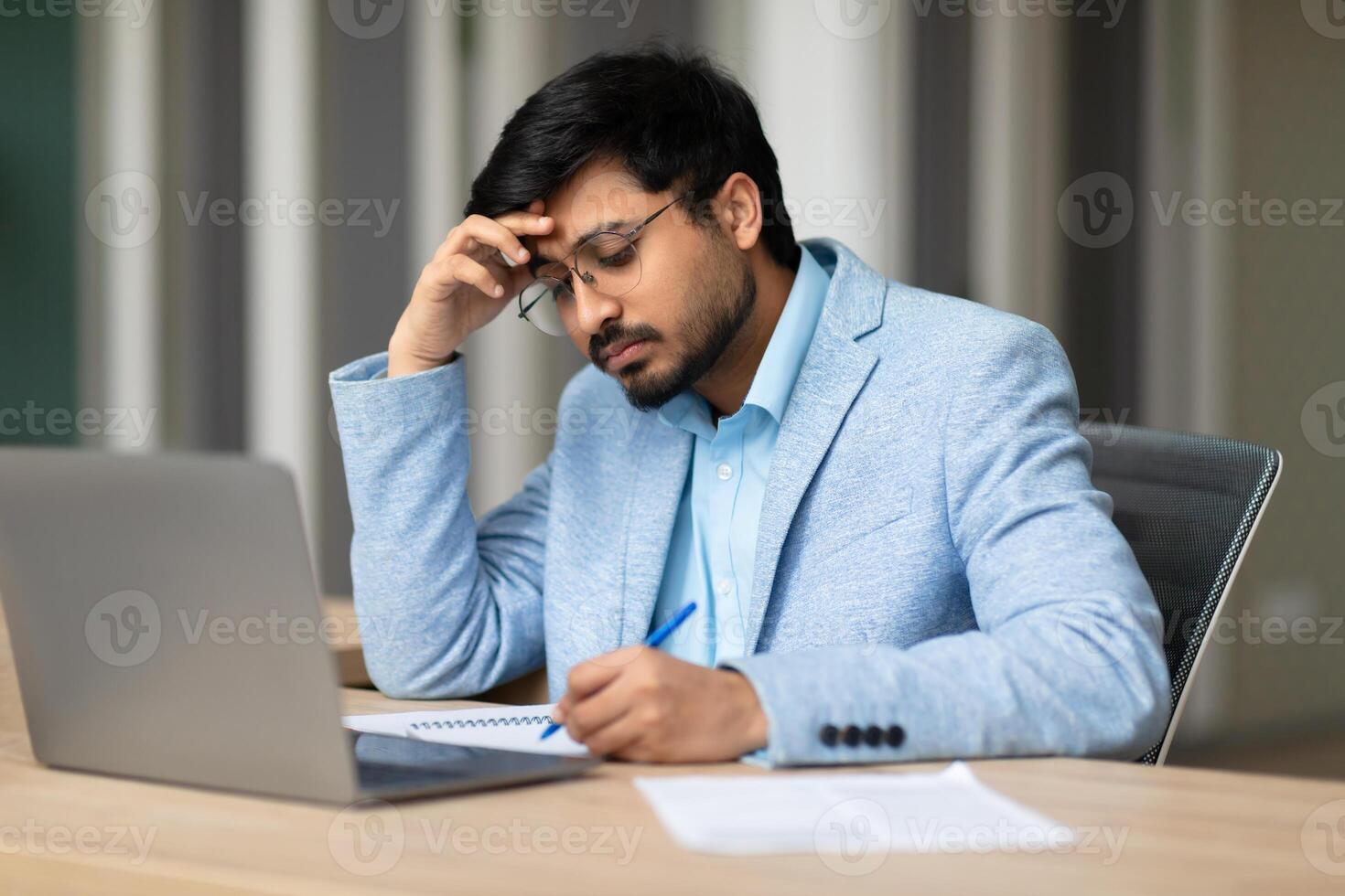Indian young man with a beard is sitting at a desk, wearing a light blue blazer and glasses, focusing intently as he works. He has his hand on his forehead and is writing on a paper with a pen photo