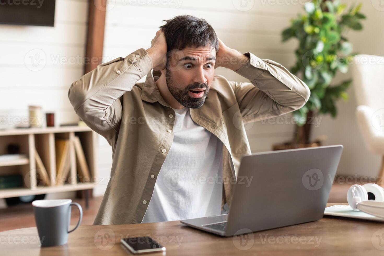 A man is seated at a wood table, hands on his head with an expression of shock and disbelief as he looks at his laptop screen. photo