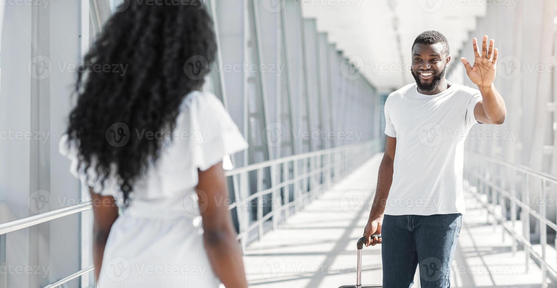 African American man with a suitcase is greeting a woman at the airport. The man is smiling and waving his hand, while the woman looks away from him. photo