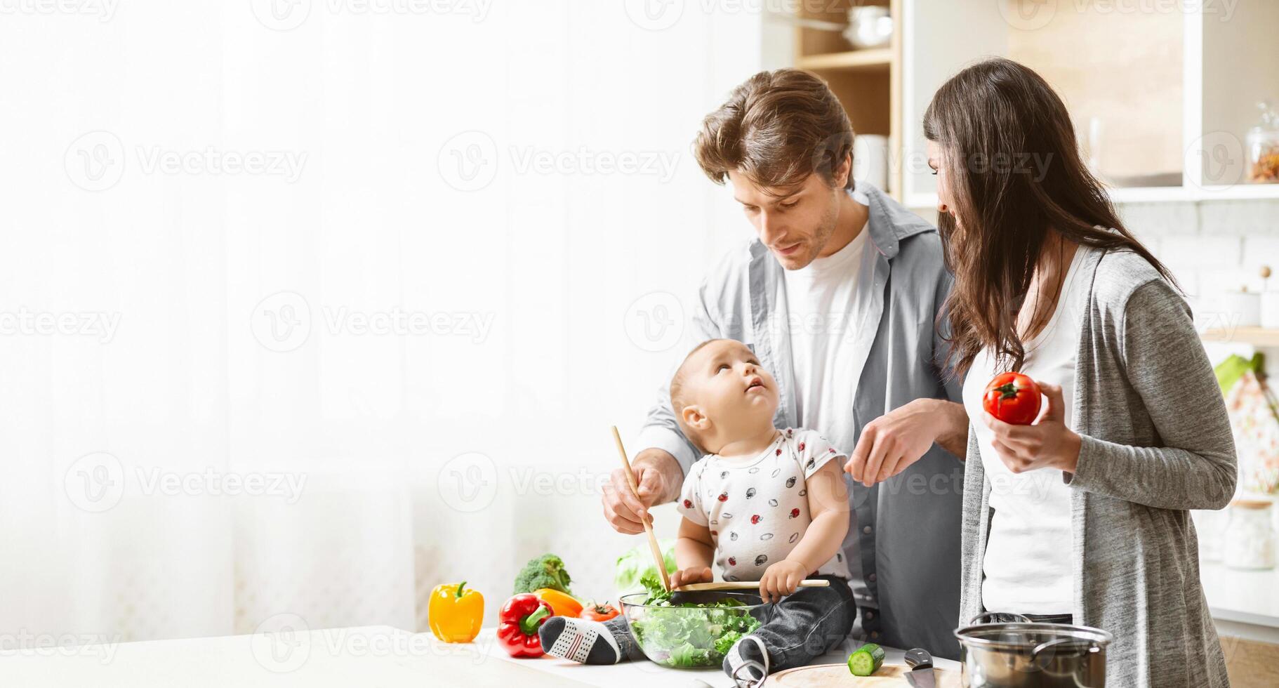 A family is cooking together in the kitchen. The father is holding a baby who is stirring a bowl of salad with a wooden spoon. The mother is standing next to them, holding a tomato, copy space photo