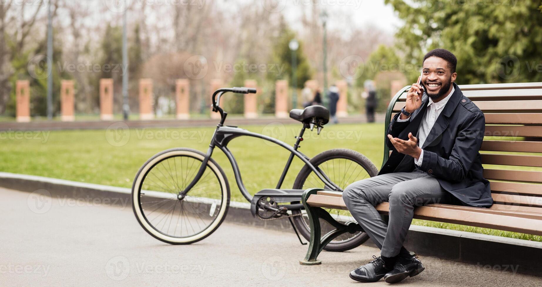 An African American man is talking on his phone in a park, sitting on a bench with his bike nearby. The setting is peaceful and green, reflecting a moment of productivity in a natural environment. photo