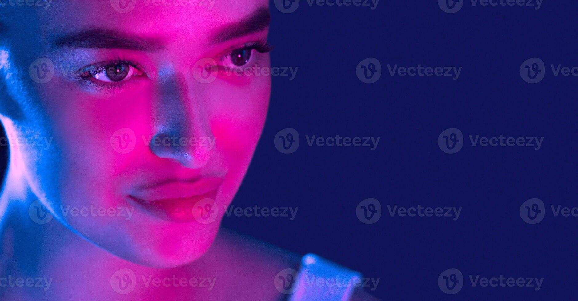 A woman face is brightly lit up with a combination of vibrant blue and pink lights, creating a striking contrast against her features. The blue and pink hues interplay on her skin, copy space photo