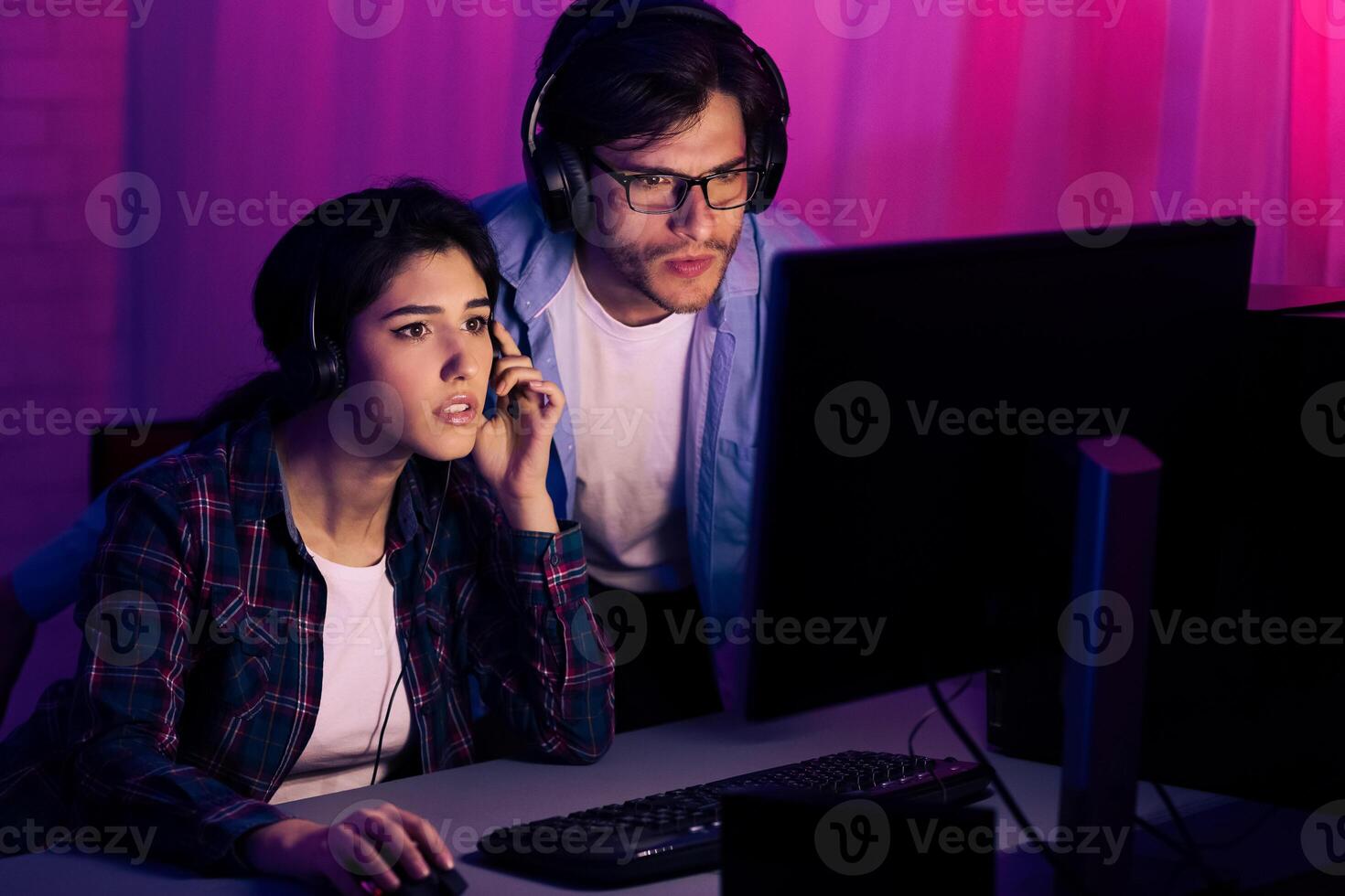 A young couple, both wearing headphones, sit in front of a computer screen in a dimly lit room. The man is standing behind the woman, both looking intently at the screen, likely engaged in a game. photo