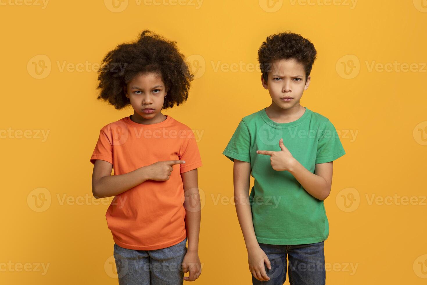 African American boy and girl, presumably siblings, stand side by side with puzzled looks on their faces, each pointing at the other against a solid yellow backdrop photo