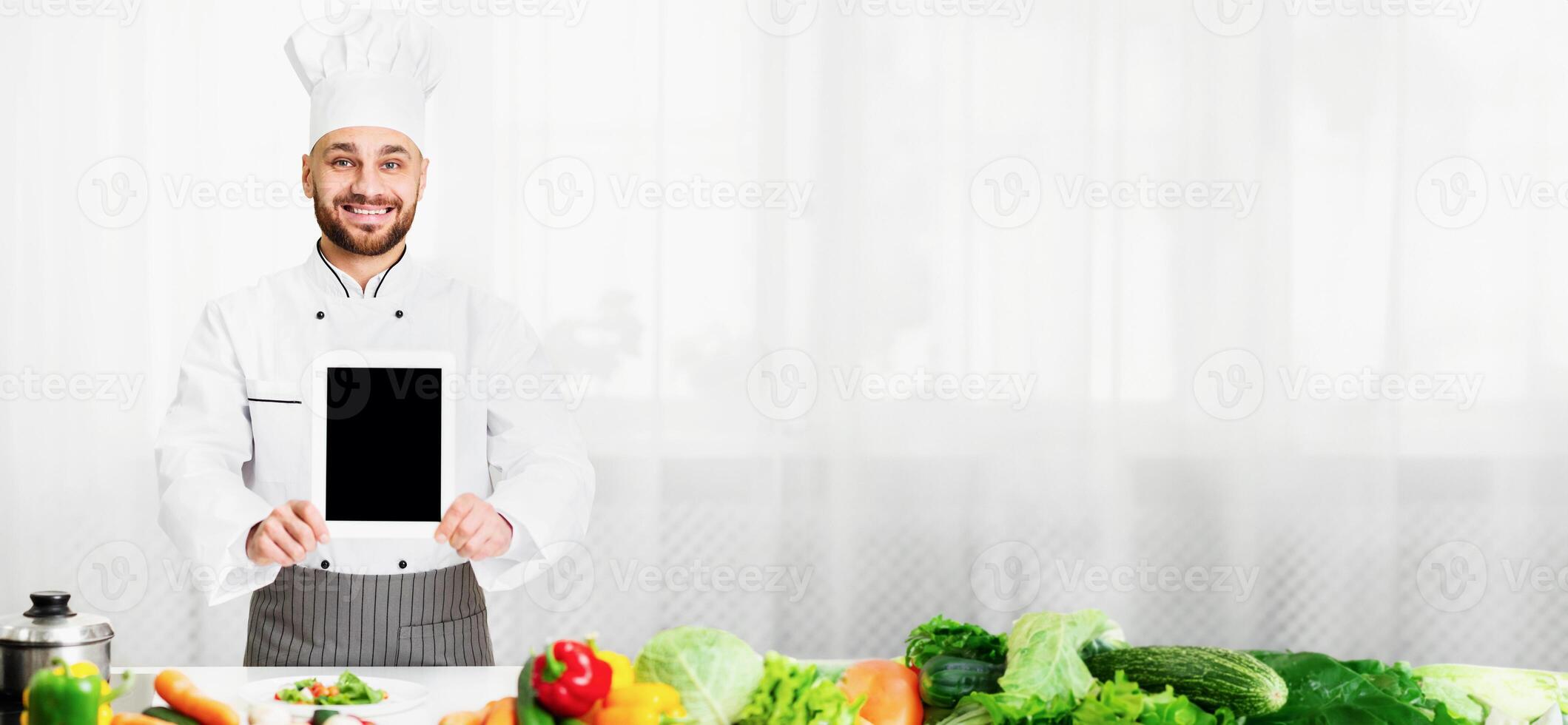 Cooking App. Chef Man Showing Digital Tablet With Empty Screen Advertising Menu Or Restaurant Application Standing In Kitchen Indoor. Mockup photo