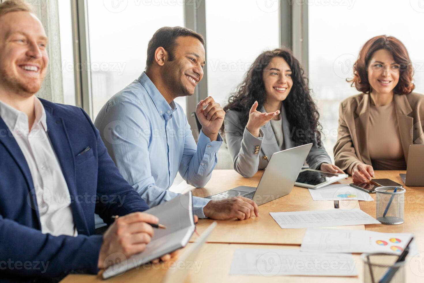 A group of business professionals, including two men and two women, participate in a meeting around a long wooden conference table. There are multiple laptops, papers, and tablets present photo