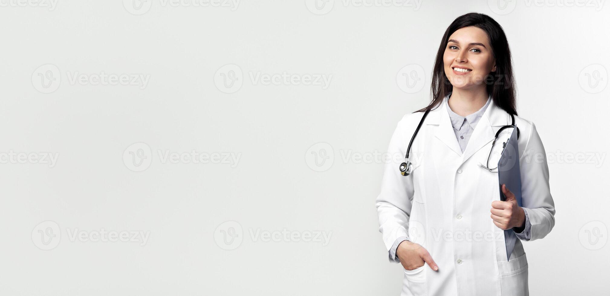 A female doctor in a white coat smiles while holding a clipboard and stethoscope. photo