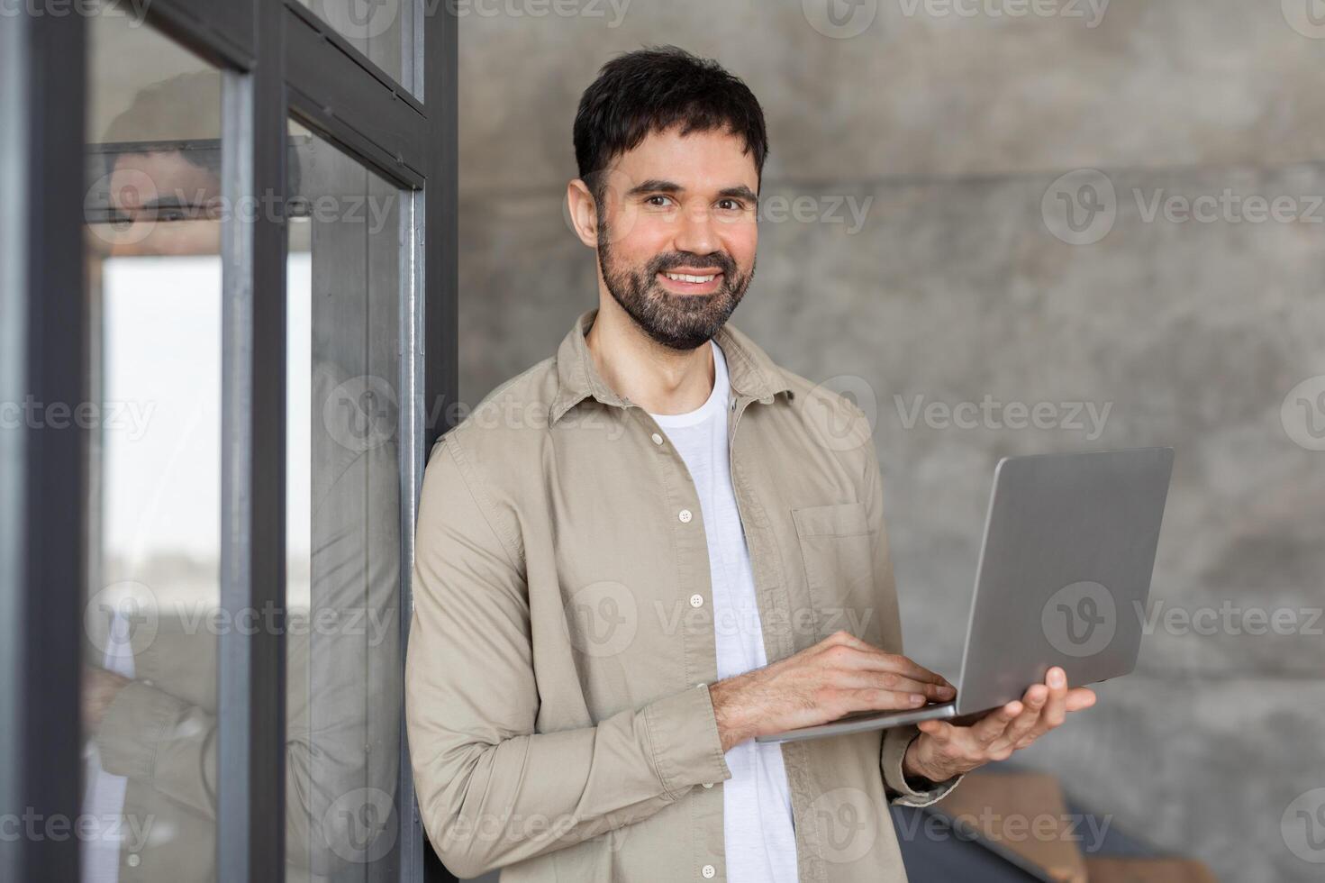 A man with short brown hair and a beard is standing in a modern office building. He is smiling and holding a laptop in his hands. photo