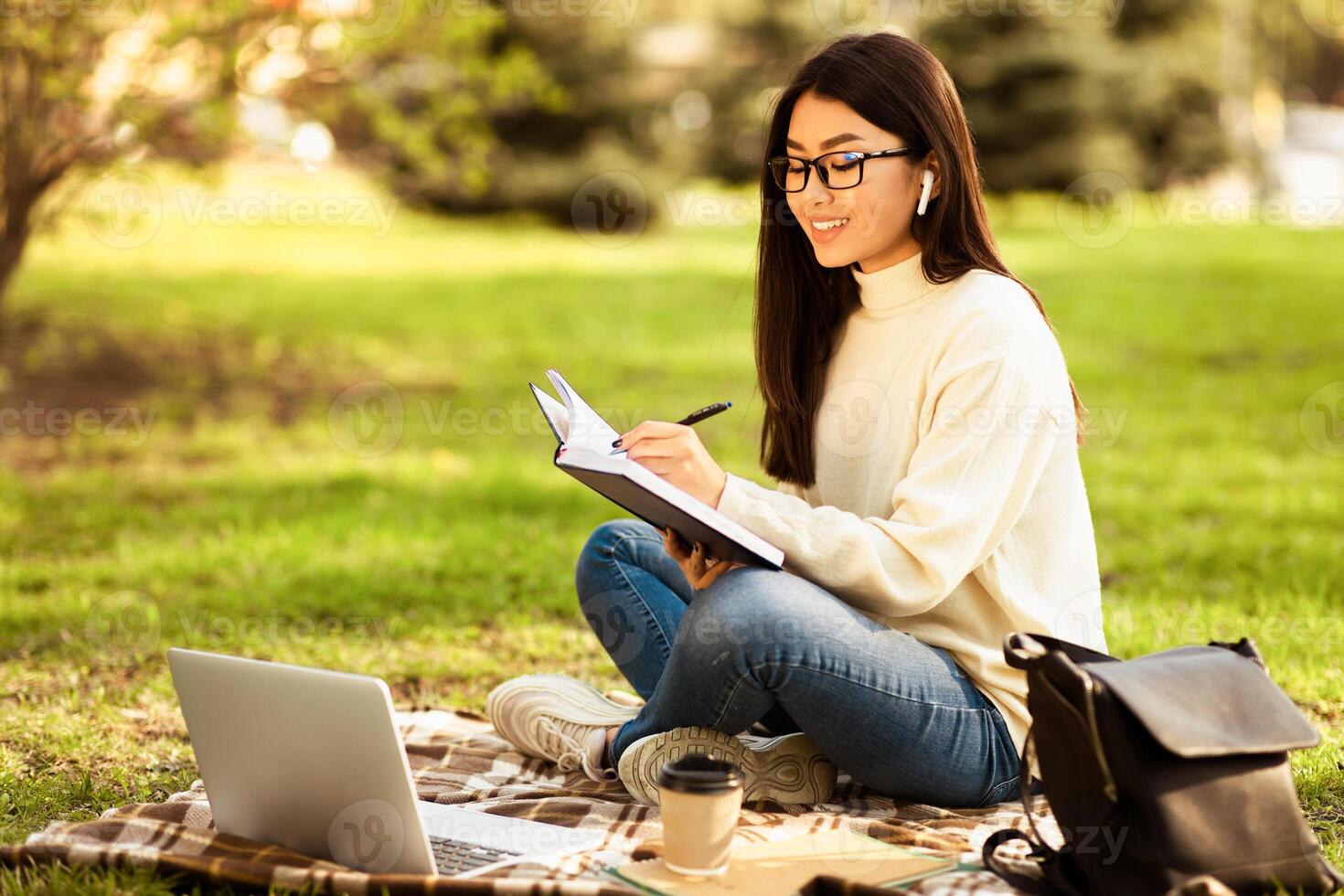 Asian woman is seated on a blanket outdoors, with a notebook and laptop open in front of her. She appears to be working or studying in a relaxed outdoor setting. photo