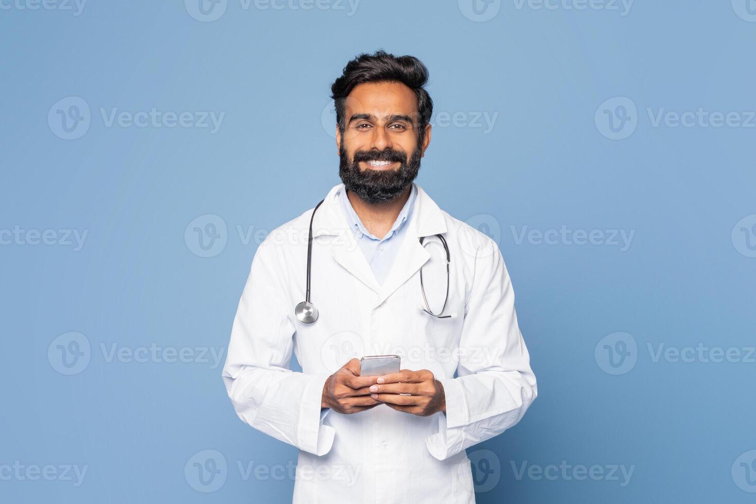 A cheerful Indian doctor in a white lab coat and stethoscope stands against a blue background, smiling as he interacts with his smartphone photo