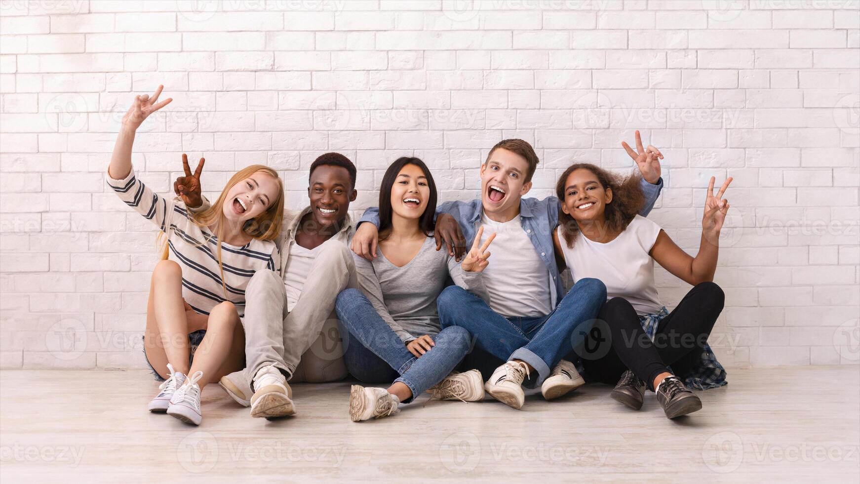Emotional multiracial students sitting on floor, showing peace gesture, posing at camera, white wall background photo