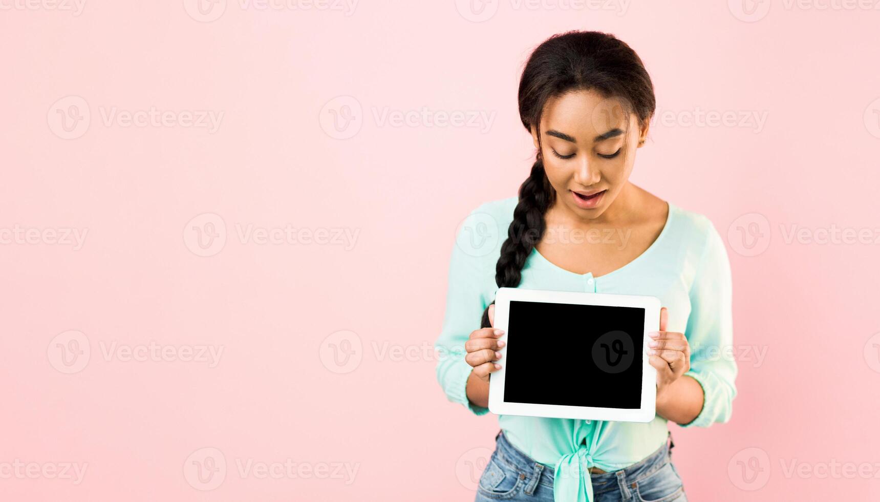 A young black girl stands against a solid pink background during the day, holding a blank tablet in both hands while looking at it, mockup, copy space photo