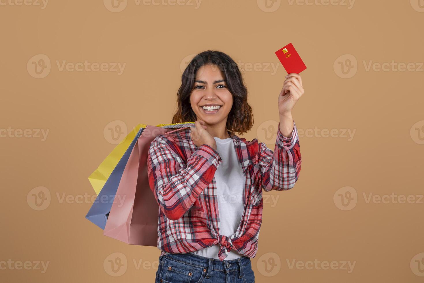 A young middle eastern woman with dark hair and a bright smile is holding a red credit card in her right hand. She is also carrying a set of colorful shopping bags over her left shoulder photo
