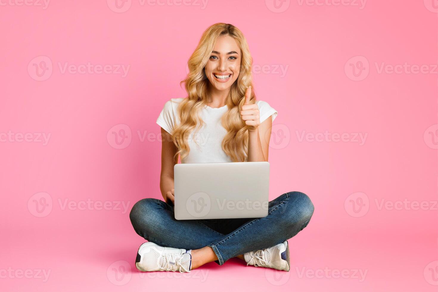 A happy woman with long blonde hair sits on the floor with her legs crossed in front of a pink background. She is using a laptop computer and giving a thumbs up with her right hand. photo