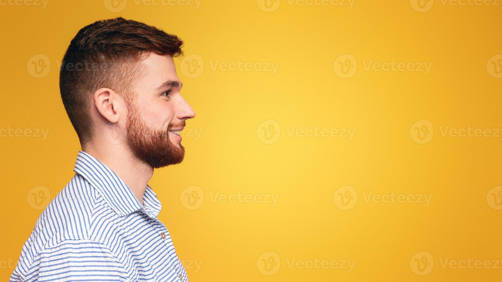 Handsome young man with a beard wearing a striped shirt posing isolated on yellow studio background, profile view, copy space photo
