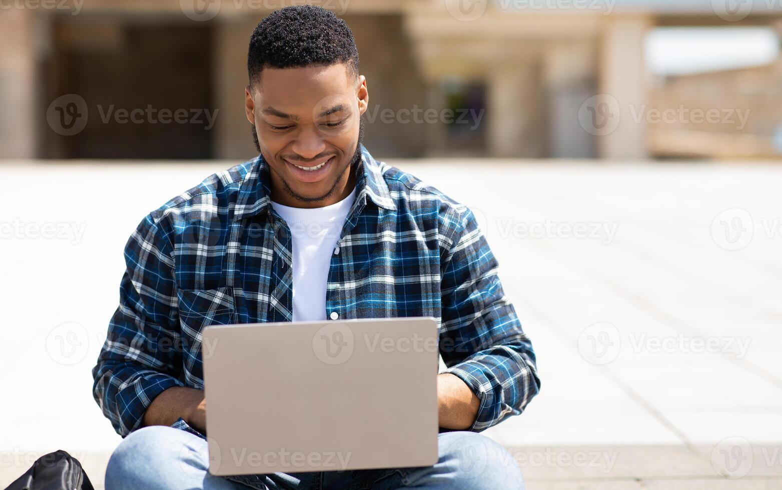 Surfing Internet. Front view portrait of smiling black person typing on laptop keyboard, sitting outdoor in park photo