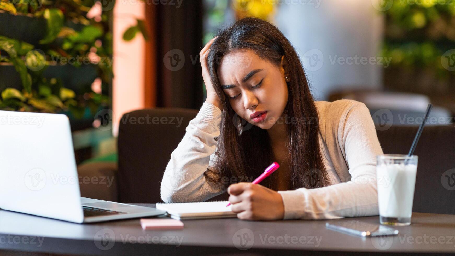 A young black girl is concentrating hard while studying in a cozy cafe. She is surrounded by a laptop, a notebook, and a pen, and appears deep in thought photo