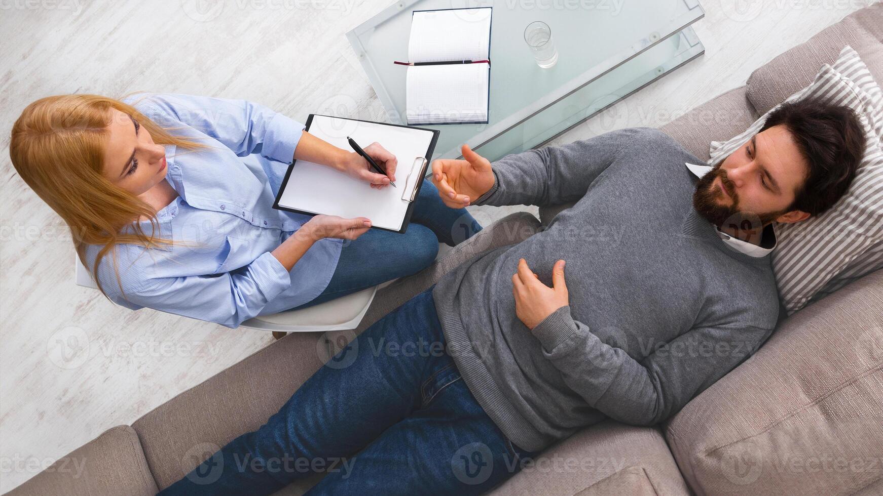 Top view of man client is reclining on a couch, discussing his thoughts with a woman therapist who is taking notes. The setting appears comfortable and private photo