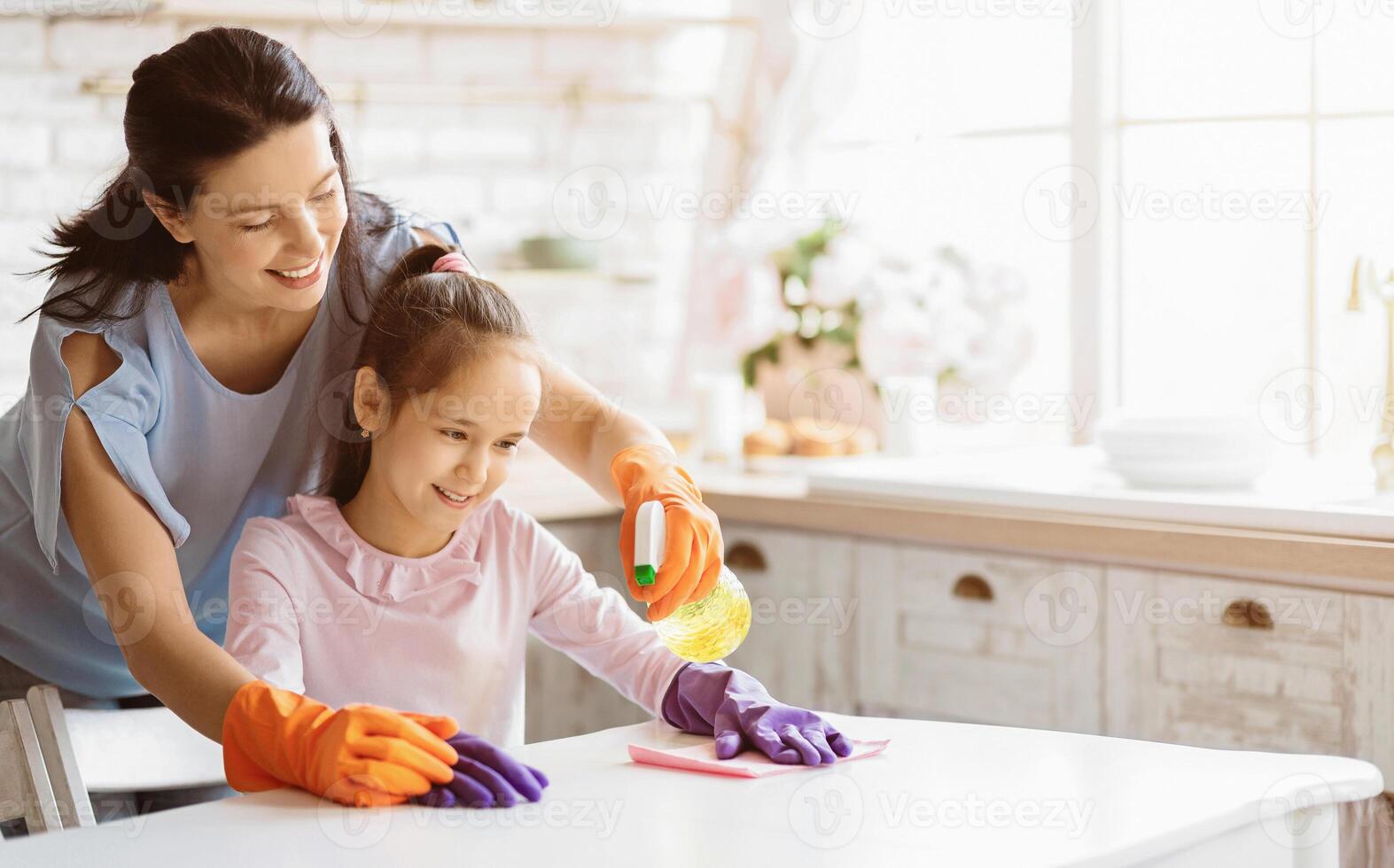 A mother and daughter are cleaning a kitchen table together. The mother is wearing orange gloves and the daughter is wearing purple gloves. They are both smiling as they work together, copy space photo