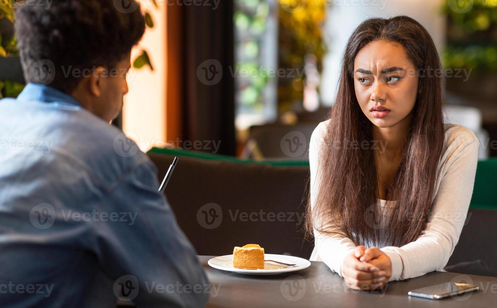 African American young girl with long hair and a white shirt sits at a table, looking concerned, while a guy in a blue shirt talks to her in a modern cafe. photo